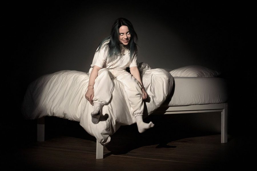 Eilish sits on a bed with light focused on her as her album cover for “When We All Fall Asleep Where Do We Go?”
