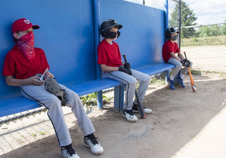 Youth baseball players, some with masks on, sit far apart as they wait for their turn to step into the batter’s box.