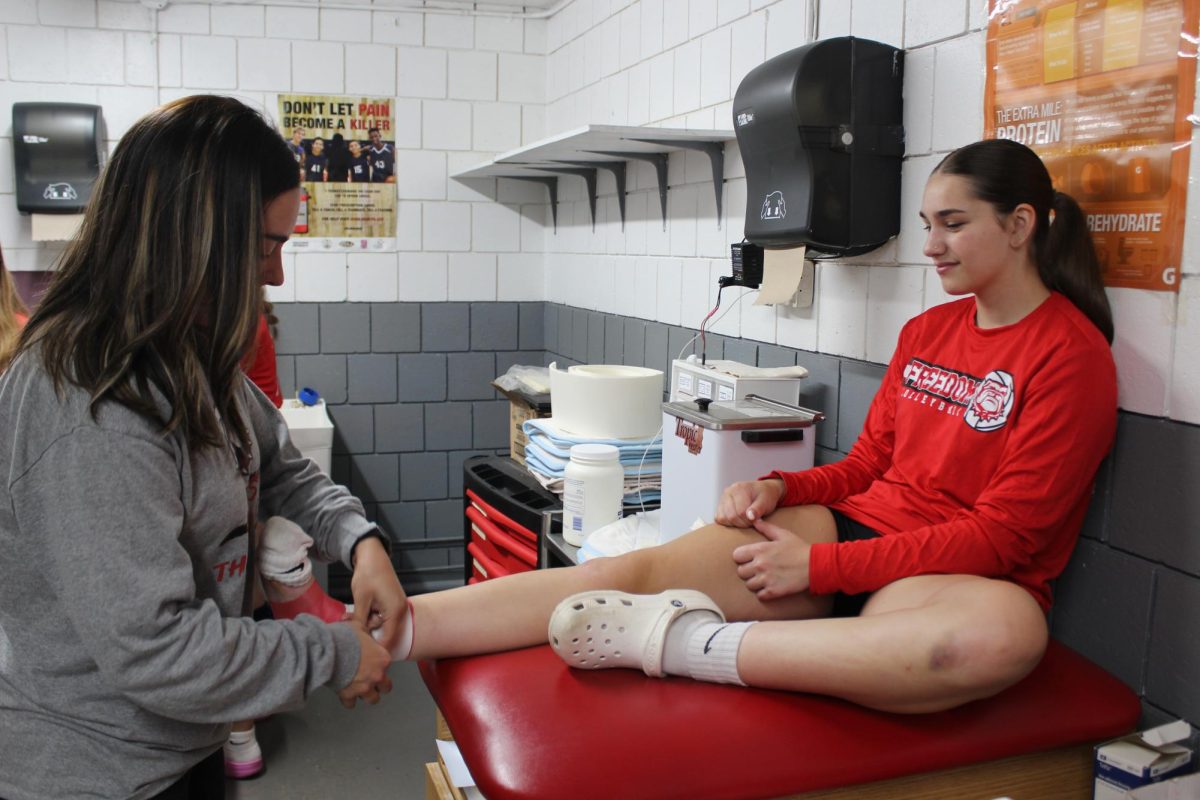 Wrapping things up: Before a big game, Alex Rawding wraps up Sophomore Riley Beegle’s ankle before beginning warm ups. Wrapping up her ankle gives her the support that she needs to allow stress on it.  
