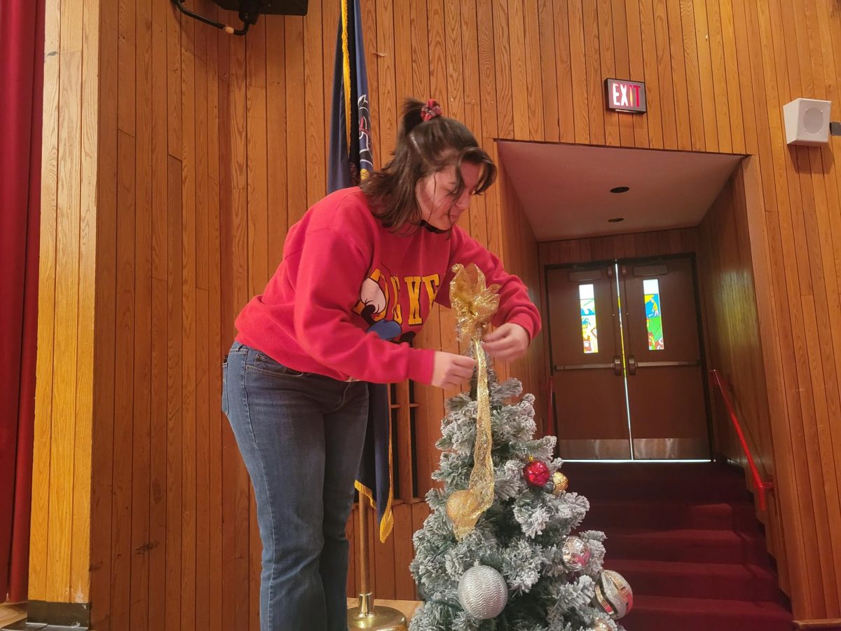 Festive and fun: Focusing, senior Lilly Burgess adjusts a gold bow atop of a Christmas tree in the high school auditorium. Burgess plays flute in the jazz ensemble, so she dedicated a portion of her practice time on Dec. 14 to decorating the tree.